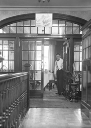 Waiter welcoming into the Luncheon Rooms inside the Carlton Restaurant, with sign pointing to Cocktail Bar upstairs. The cocktail bar opened in May 1936. * 