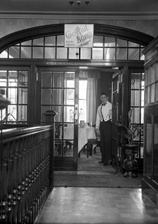 Waiter welcoming into the Luncheon Rooms inside the Carlton Restaurant, with sign pointing to Cocktail Bar upstairs. The cocktail bar opened in May 1936. * 
