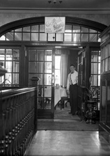 Waiter welcoming into the Luncheon Rooms inside the Carlton Restaurant, with sign pointing to Cocktail Bar upstairs. The cocktail bar opened in May 1936. * 