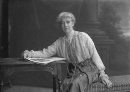 Elderly woman leaning on desk reading a book, wearing satin skirt with timepiece on a long cord.#