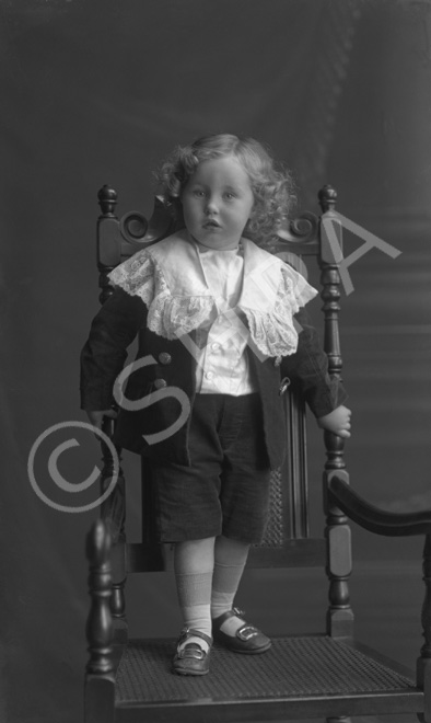 Male child with long blonde ringlets standing on a chair wearing a Little Lord Fauntleroy suit.#