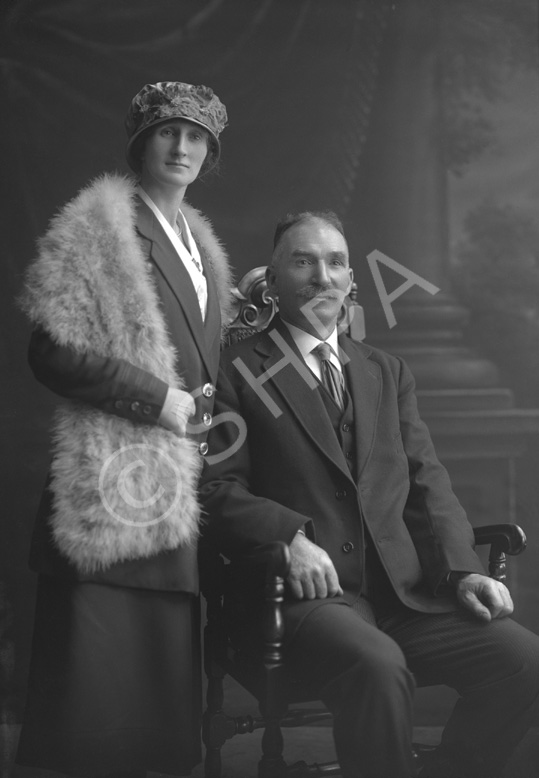 Couple, he seated wearing suit, she standing wearing hat and fur shawl.#.....