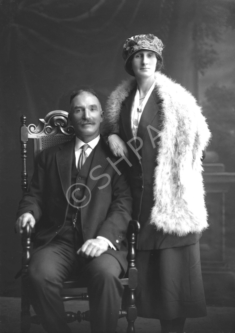 Couple, he seated wearing suit, she standing wearing hat and fur shawl. #.....